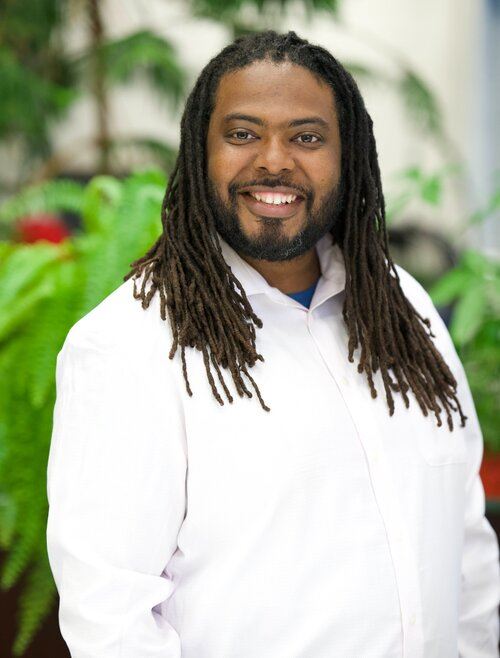 Headshot of an African American man with a beard and long locs. He is wearing a white shirt and stands against green ferns in the background.